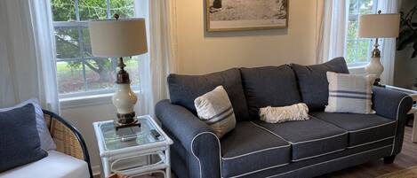 A living room with a dark grey sofa, white cushions, a glass coffee table, and a framed picture on the wall.