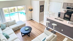 A modern living room with a sofa set, a coffee table, and a dining area with a table and chairs. A kitchen with appliances is visible in the background.