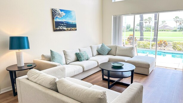 A modern living room with a sectional sofa, a round coffee table, and a lamp. There is a painting on the wall and a view of a pool through large windows.