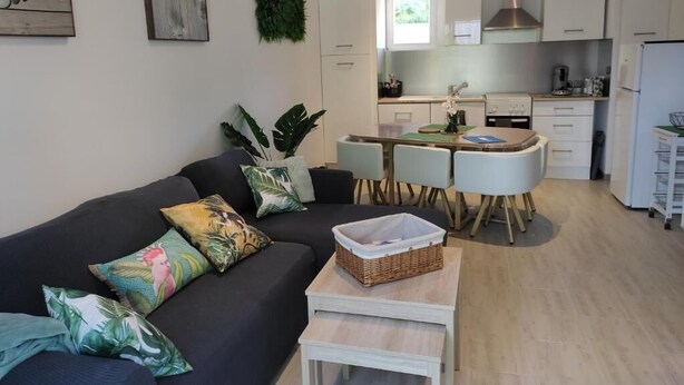 Ein modernes Wohnzimmer mit einem dunklen Sofa, einem kleinen Couchtisch und einem Essbereich mit weißem Tisch und Stühlen. Im Hintergrund ist eine Küche mit weißen Schränken und einer Edelstahl-Dunstabzugshaube zu sehen.