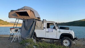 En vit terrängbil med ett stort campingtält fastsatt på bakpartiet, parkerad vid en sjö under en klar himmel.
