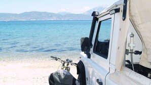 En vit bil står parkerad vid en strand med fri sikt över havet och de avlägsna bergen.