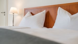 Un letto ben rifatto con lenzuola e cuscini bianchi, una testiera in legno e una lampada da comodino.