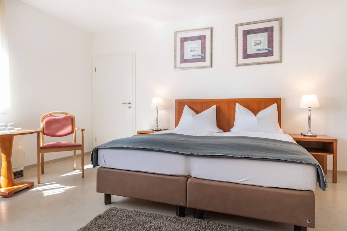 Una cama doble con cabecera de madera, dos mesitas de noche con lámparas, una silla roja y dos cuadros enmarcados en la pared.