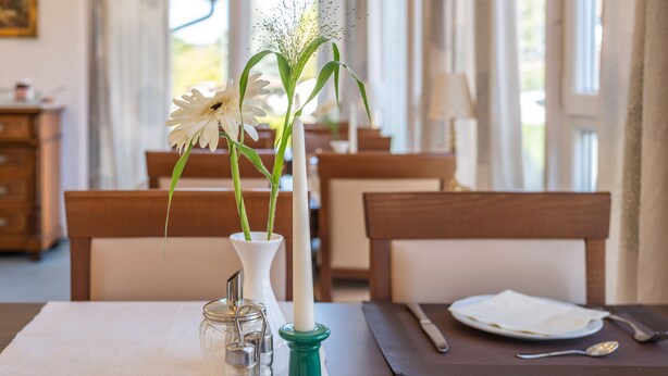 Un tavolo da pranzo con un vaso bianco pieno di fiori bianchi, una candela bianca alta e un portacandele verde.