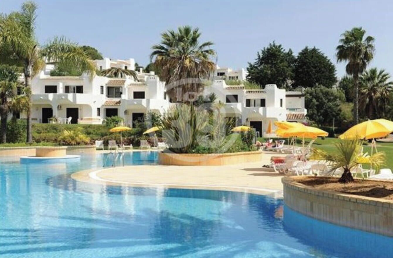 Piscina con sillones de descanso y sombrillas, rodeada por un edificio blanco con balcones.
