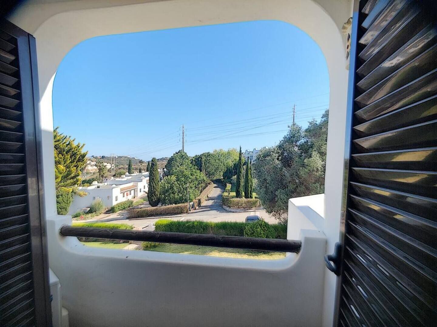 Vista a través de una ventana con persianas, que muestra una zona residencial con casas, árboles y cielo despejado.