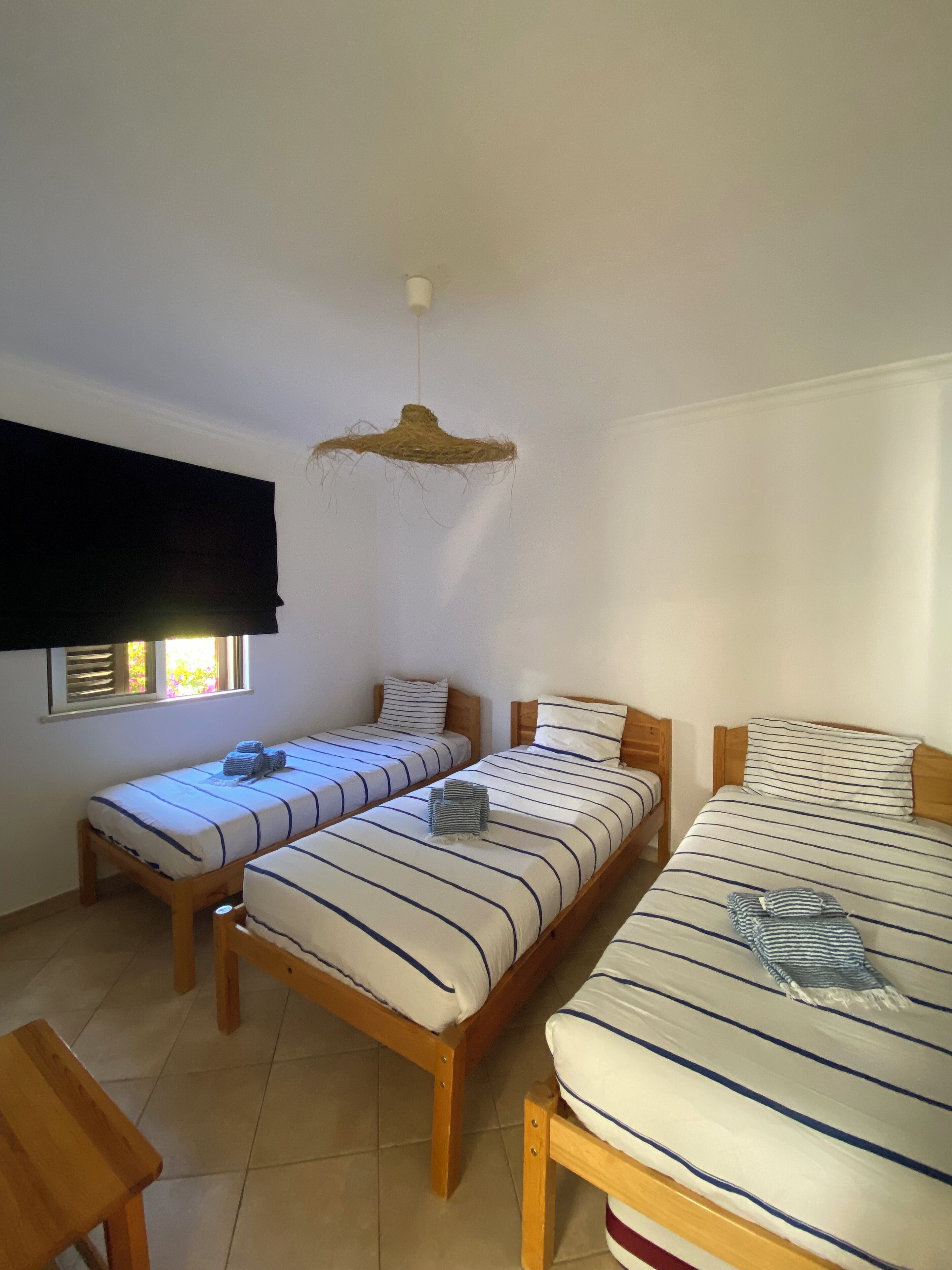 Une chambre avec trois lits simples, une table en bois, un luminaire suspendu et une fenêtre avec des stores.