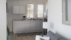 Ein modernes Wohnzimmer mit einem grauen Sofa, gefliestem Boden und einer Küche im Hintergrund mit weißen Schränken und einem Fenster mit Jalousien.