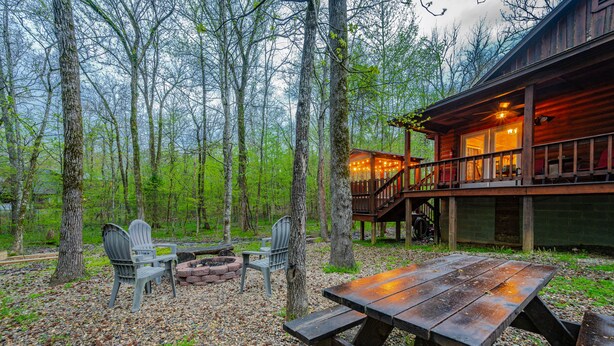 Une cabane en bois dotée d’une terrasse et d’un foyer extérieur, entourée d’arbres.