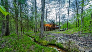 Un chalet rustique, entouré d’une forêt dense, avec un petit ruisseau qui le traverse.