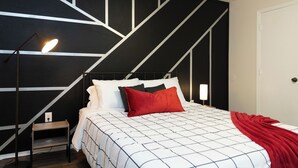 Un dormitorio moderno con un diseño geométrico en la pared, una cama bien tendida con un almohadón rojo y una lámpara de noche.