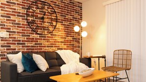 Una sala moderna con pared de ladrillo, un sofá negro con cojines, una mesa de centro de madera, una silla de mimbre y un reloj de pared redondo.