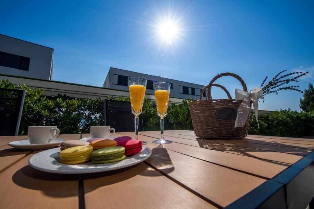 A table with a plate of colorful macarons, two glasses of orange juice, and a white cup on a sunny day.