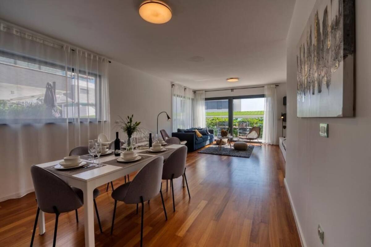 A modern dining area with a white table set for four, grey chairs, and a large window with sheer curtains.