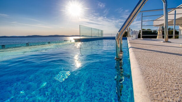 An infinity pool with clear blue water, a metal handrail, and a view of the sea and distant hills under a bright sun. - Crystal Holiday Residence (Sukosan)