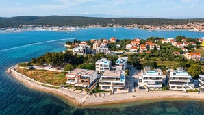 An aerial view of a coastal area with modern buildings, a beach, and a clear blue sea. - Crystal Holiday Residence (Sukosan)