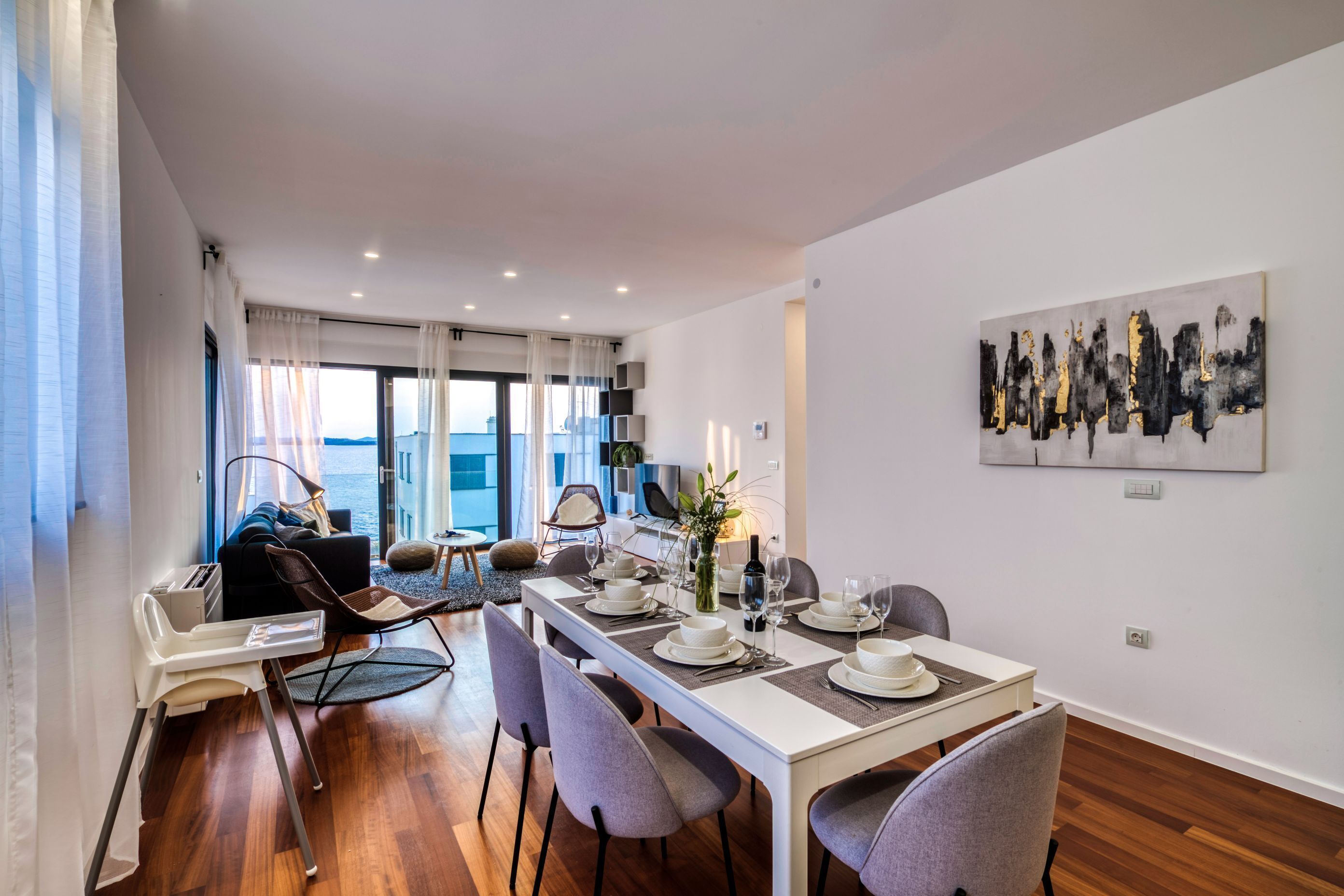 A modern dining area with a long table set for four, surrounded by grey chairs. A white high chair is positioned to the left, and a sofa with a view of the sea is in the background.