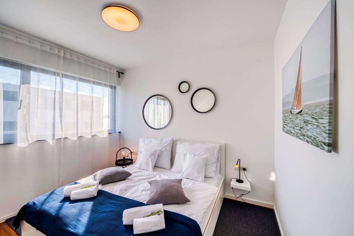 A modern hotel room with a neatly made bed, a bedside table with a lamp, and a large window with sheer curtains.