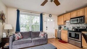 Ein kompakter Wohnraum mit grauem Sofa, einem Esstisch, einer Küchenzeile mit Mikrowelle und Herd sowie einem Fenster mit Vorhängen.