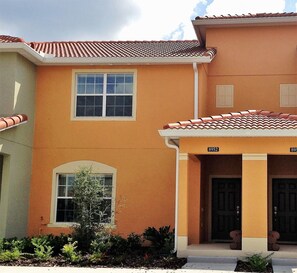 A two-story house with a tiled roof, a front porch, and a small garden area. - Moderno Townhouse 4 hab· Piscina · Resort con Cine & Tobogán, 8 millas de Disney (Kissimmee)