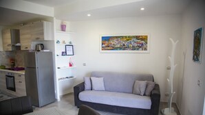 A modern living room with a grey sofa, a dining table with chairs, a kitchen area with a refrigerator and stove, and a painting of a coastal village on the wall. - Nice apartment not far from the sea (Lacco Ameno)