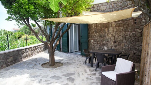 An outdoor patio with stone flooring, a table with chairs, a wicker sofa, and a large tree providing shade. - Nice apartment not far from the sea (Lacco Ameno)