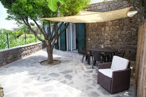 An outdoor patio with stone flooring, a table with chairs, a wicker sofa, and a large tree providing shade. - A nice apartment located not far from the ocean (Lacco Ameno)