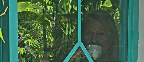 Una ventana con un diseño geométrico, una persona sosteniendo una taza y un exuberante follaje verde al fondo.