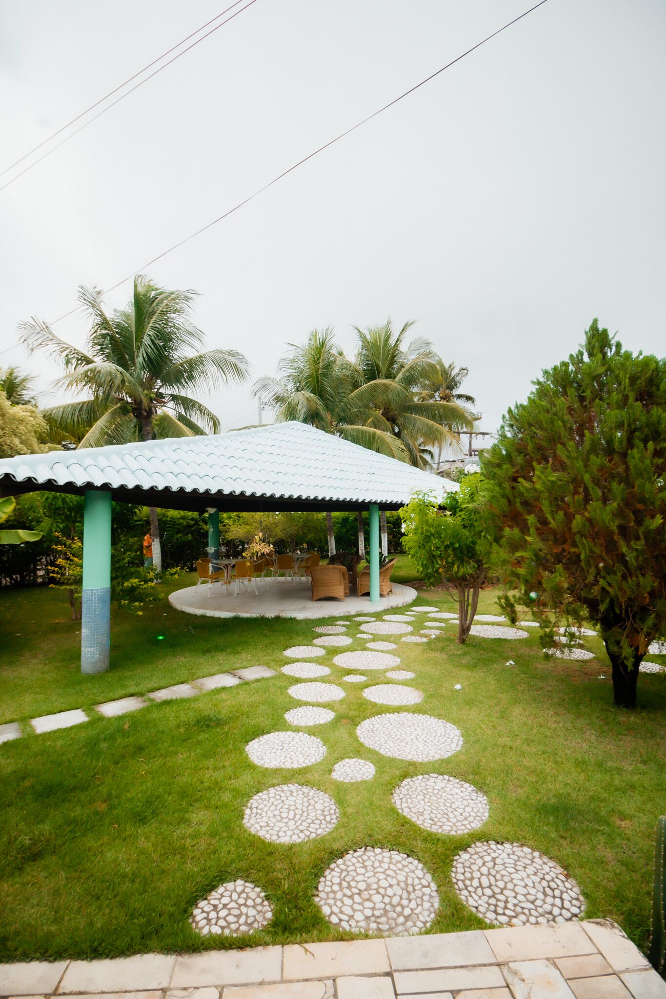 A covered outdoor seating area with a tiled roof, surrounded by palm trees and a patterned stone pathway.