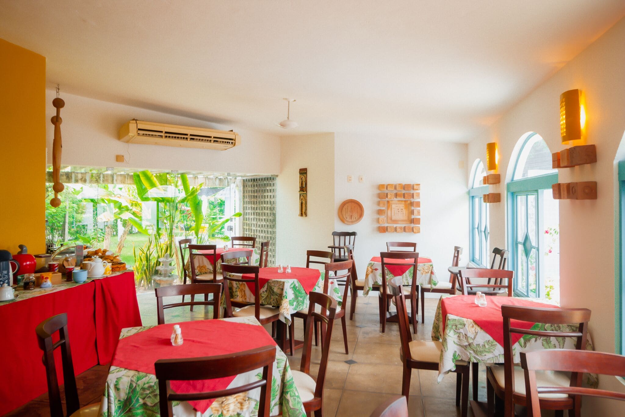 A restaurant with tables covered in red tablecloths, wooden chairs, and a view of greenery outside.