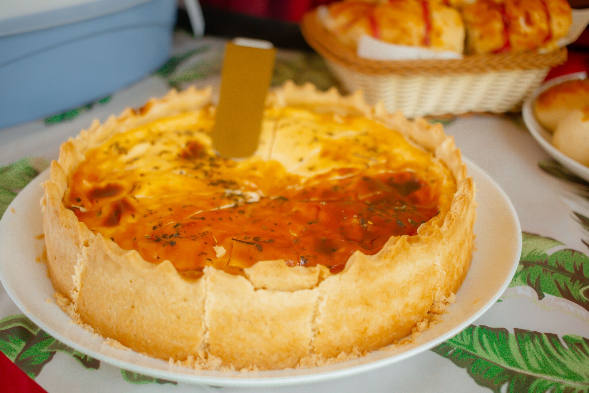 Quiche rodona sobre un plat blanc amb un ganivet al centre, damunt d'una taula amb estampa floral.