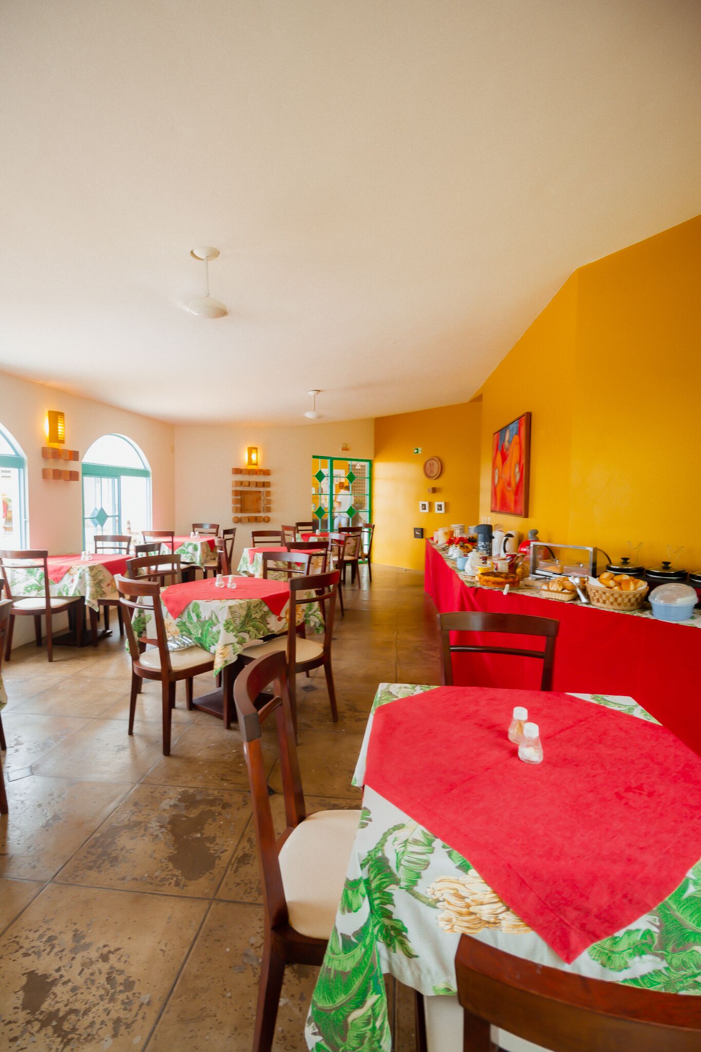 Um restaurante com mesas cobertas por toalhas vermelhas e verdes, cadeiras de madeira e uma parede amarela.