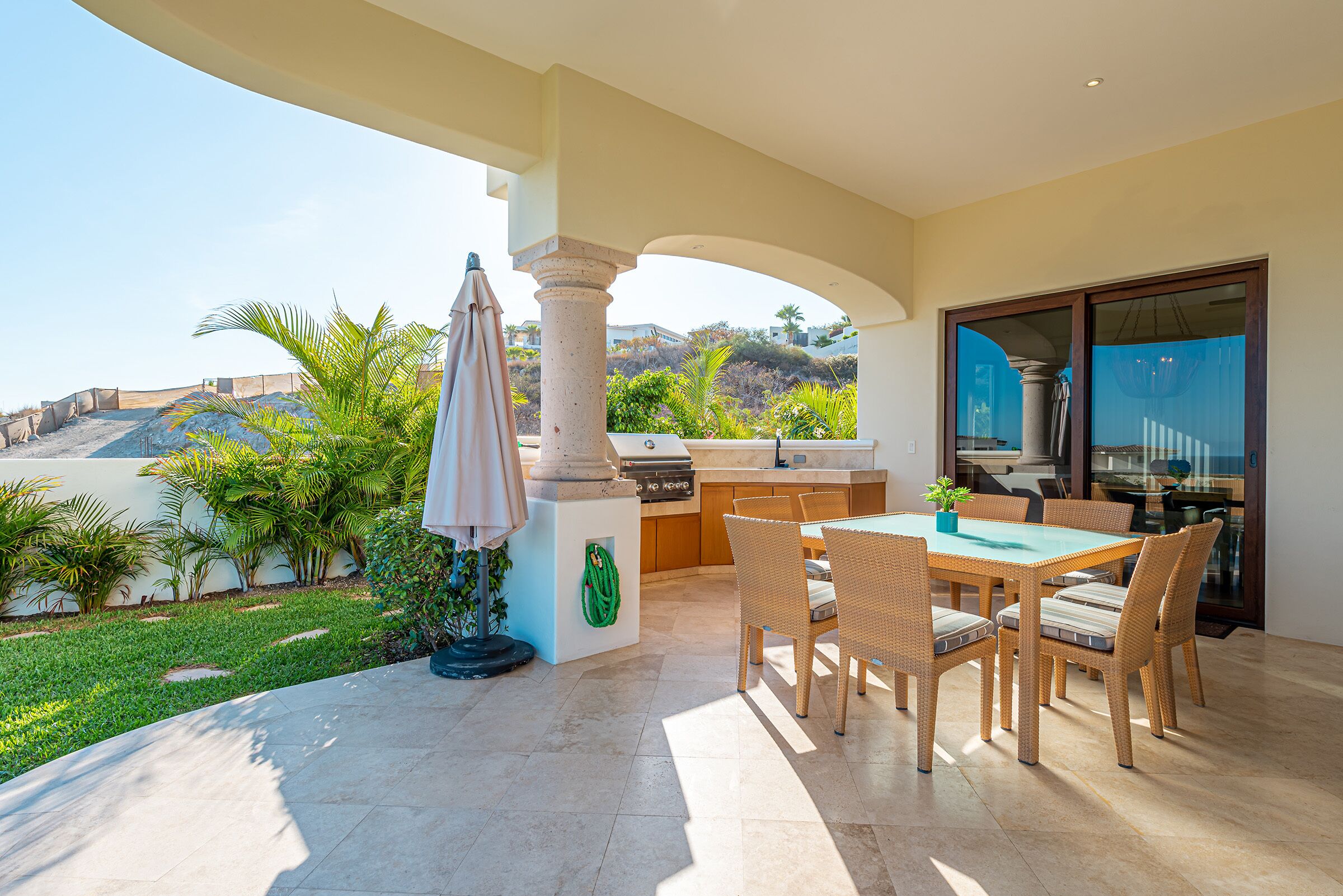 An outdoor patio with a dining table and chairs, a grill, and a view of a landscaped area with palm trees.