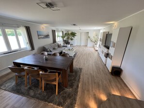 Un salon moderne avec une table à manger en bois, un canapé et des chaises. Un téléviseur est fixé au mur et une plante se trouve dans un coin.