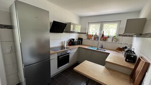 A modern kitchen with a built-in refrigerator, oven, and sink. There is a wooden table and a chair, and the kitchen has a window with a view of trees.