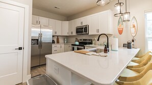 A modern kitchen with white cabinets, stainless steel appliances, a central island with seating, and decorative surfboards on the wall. - New 3 bed 4 bath home - rooftop deck, 2 car garage & porch near downtown! (Omaha)