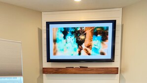 A living room with a flat-screen TV mounted above a fireplace, a yellow armchair with a cushion, a round coffee table with books, and a teal ottoman. - New 3 bed 4 bath home - rooftop deck, 2 car garage & porch near downtown! (Omaha)