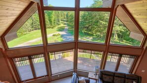 Ein Zimmer mit großem Fenster, das einen Blick auf eine grüne Landschaft und eine Holzterrasse bietet. Es gibt ein Ledersofa und einen kleinen Tisch mit einer Lampe.