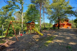 Una estructura de juego de madera con un tobogán, una casita de madera y un sendero de grava en un área boscosa.
