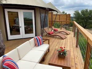 Een houten terras met een bank en twee fauteuils, een tafel met een bloemstuk, en een glazen deur naar een binnenruimte.