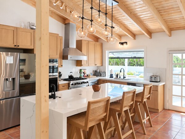 A modern kitchen with a central island, wooden cabinets, stainless steel appliances, and a large window providing natural light.