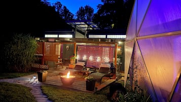 Un patio moderno al aire libre con una chimenea, asientos y una estructura con cerramientos de vidrio.