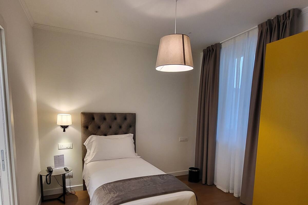 A hotel room with a bed, bedside table with a lamp, a ceiling fan, and a large window with curtains.