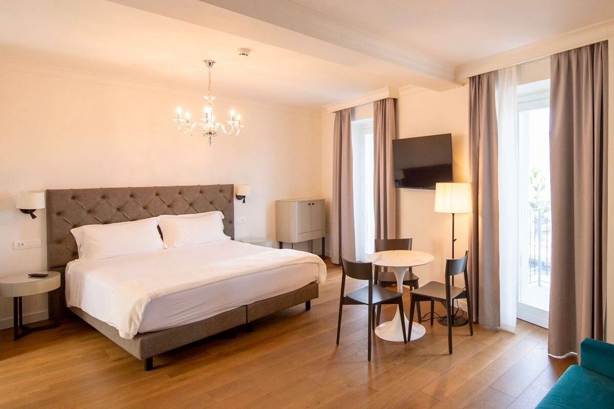 A hotel room with a large bed, a chandelier, a TV, a desk with chairs, and a sofa.