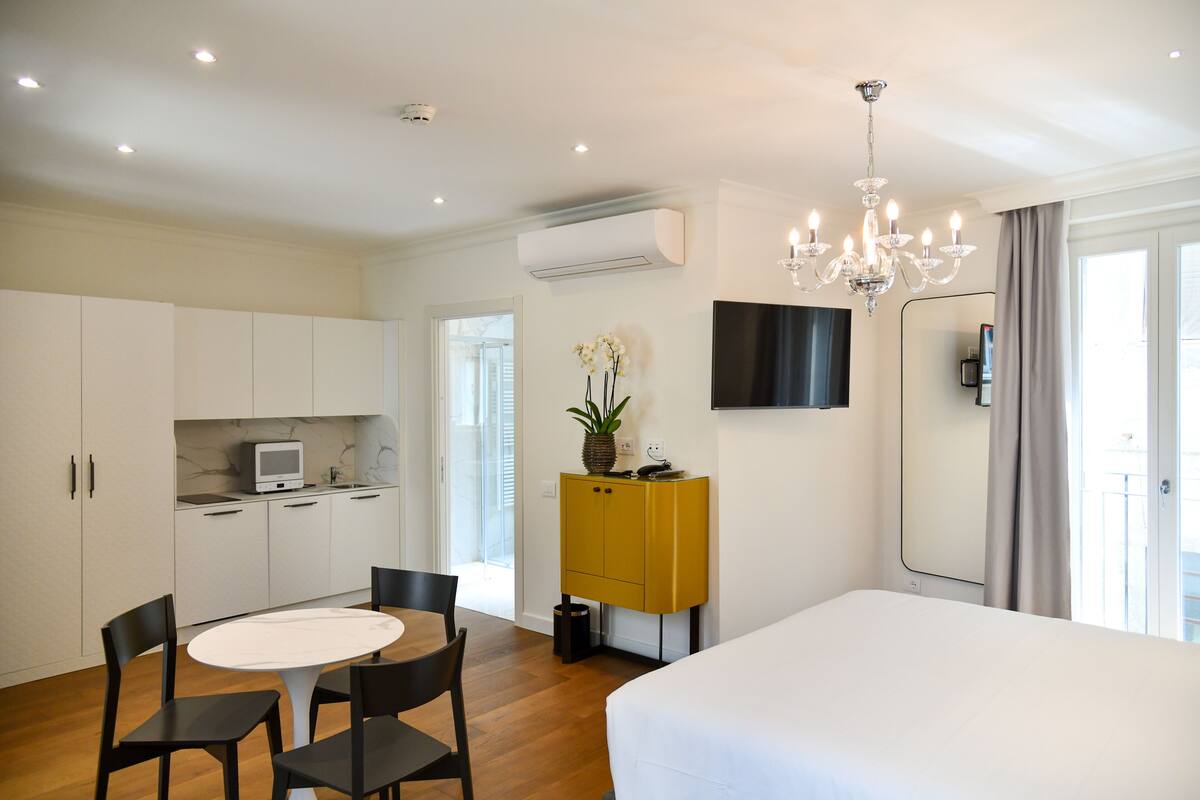A hotel room with a bed, a dining table with chairs, a small kitchen area, a TV mounted on the wall, and a large window with curtains.