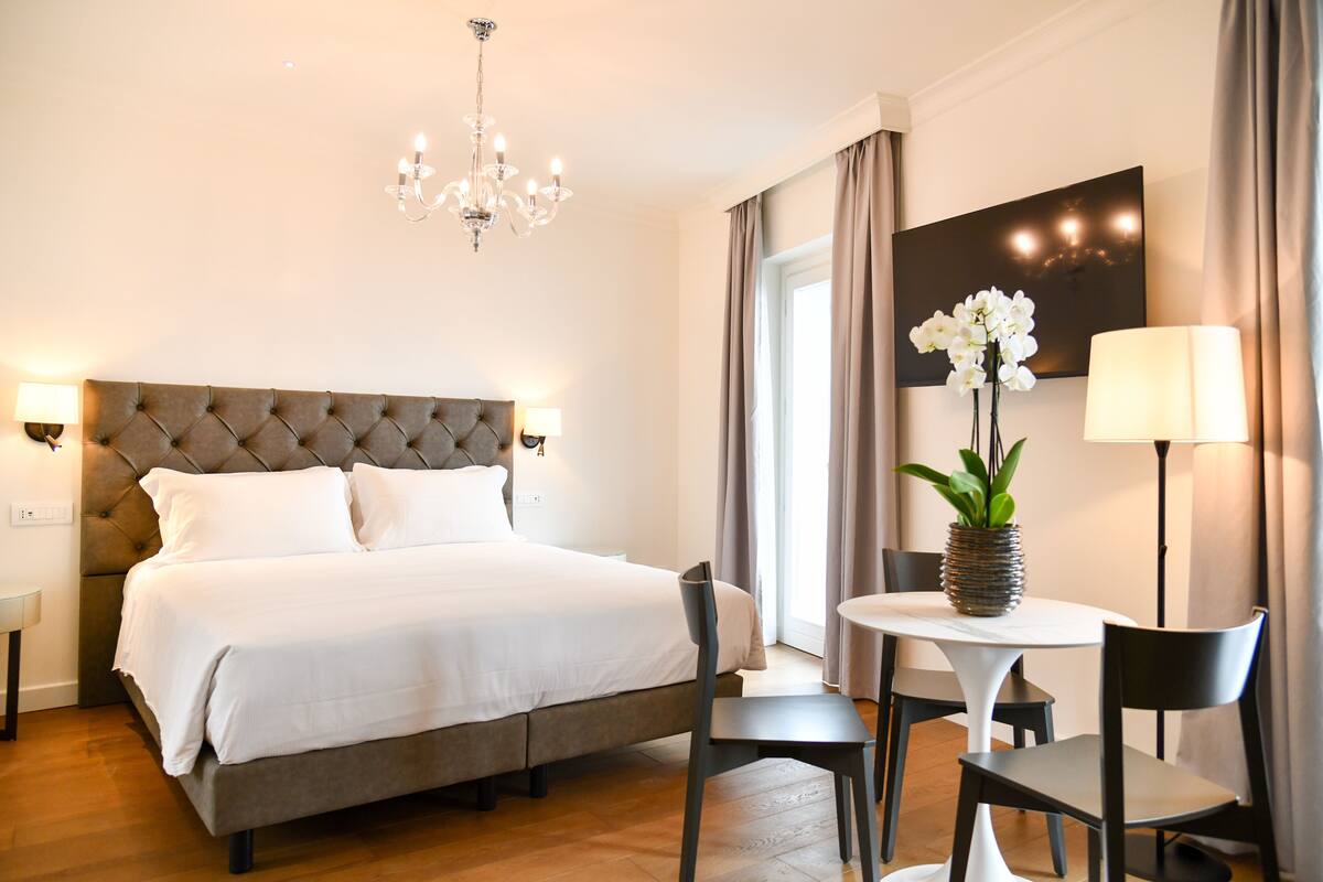 A hotel room with a large bed, a chandelier, a dining table with chairs, and a vase with flowers.