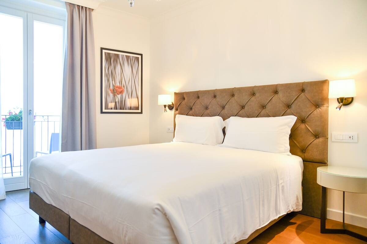 A hotel room with a large bed, a small round table, a wall-mounted lamp, and a framed picture on the wall.