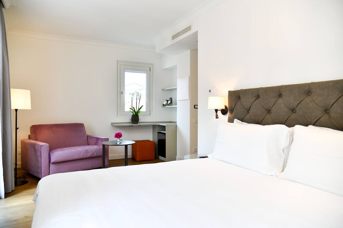 A hotel room with a large bed, a purple armchair, a small table with a vase, and a desk with a chair.