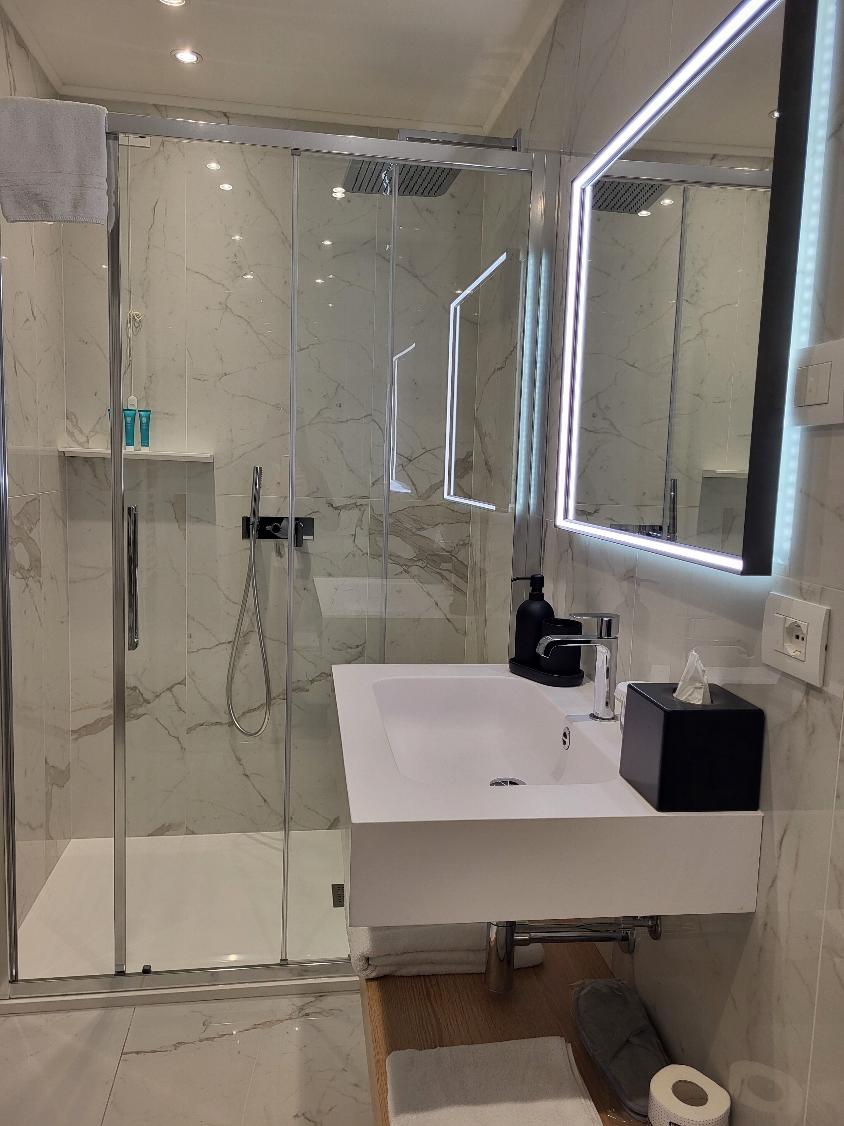 A modern bathroom with a glass-enclosed shower, a white sink with a black soap dispenser, and a large mirror.